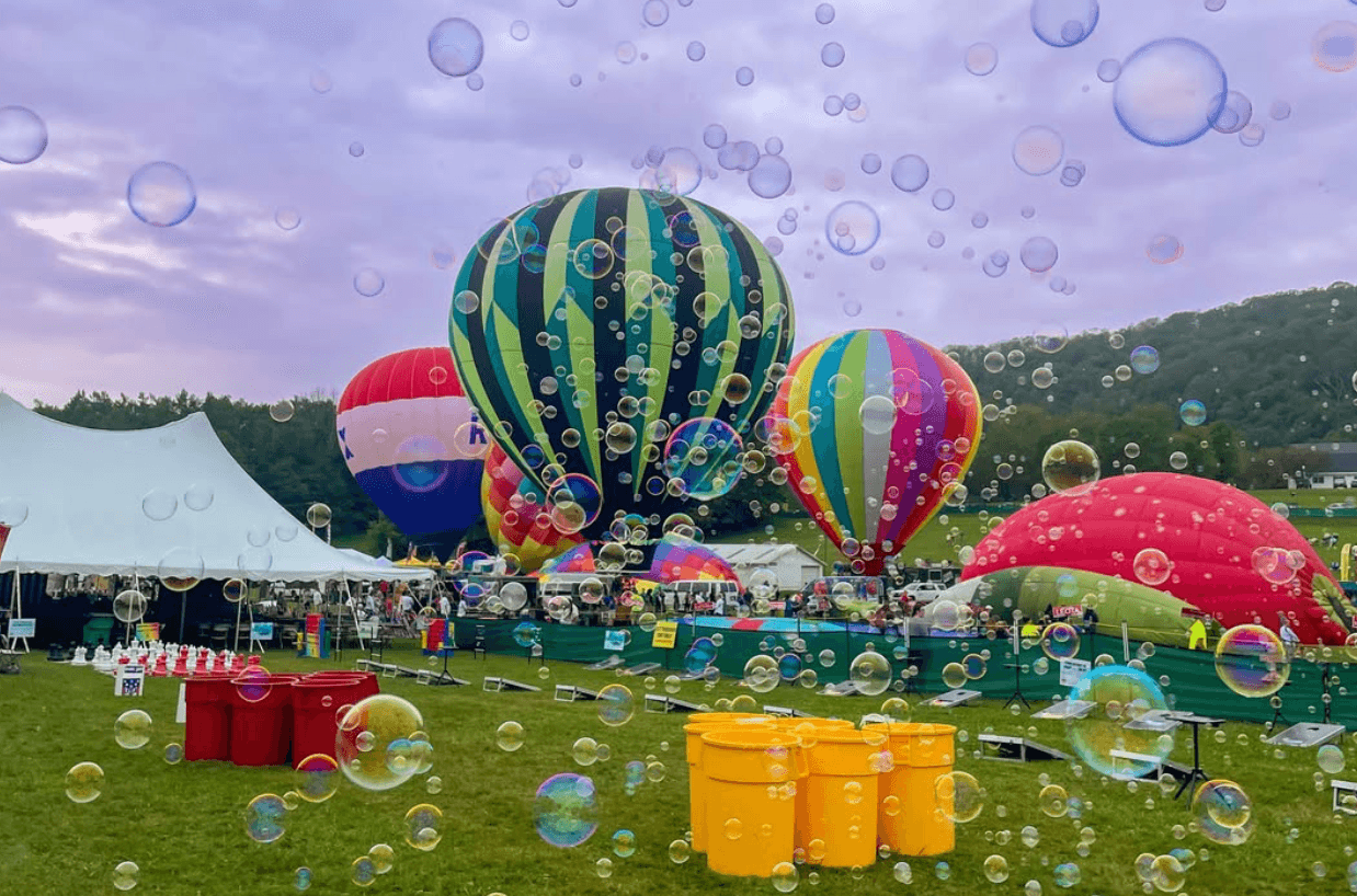 Colorful hot air balloons being inflated on a grassy field at a Hot Air Balloon Festival, with bubbles floating in the air, people gathered near tents, and trees in the background under a vibrant purple sky.